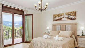 Un dormitorio con una cama grande, dos mesitas de noche, una araña, un cuadro y un balcón con vistas.