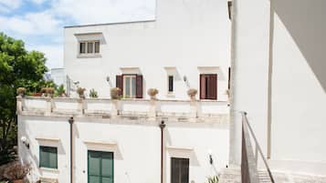 Un edificio blanco con múltiples ventanas y puertas, un balcón con plantas en macetas y un cielo despejado.