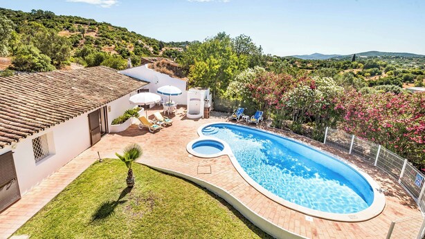 A swimming pool with a curved edge, surrounded by a brick patio and a small lawn. There are lounge chairs and umbrellas near the pool, and a white building with a tiled roof in the background. - Casa Nomanico - Two Bedroom Villa, Sleeps 4 (Estoi)