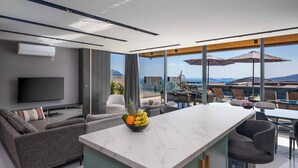 Un salon moderne doté d’un téléviseur à écran plat, d’un canapé, d’un coin repas avec des chaises et des parasols, et d’une grande fenêtre offrant une vue sur la mer.