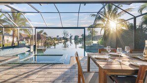 Un espace repas aménagé au bord de la piscine, offrant une vue sur un plan d’eau et des palmiers, avec une table dressée et une structure entièrement vitrée.