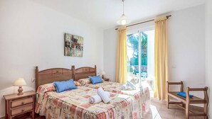 A bedroom with two beds, a nightstand with a lamp, a wooden chair, and a balcony with yellow curtains. - Villa Cati Punta Prima (Punta Prima)