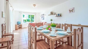 A dining area with a wooden table set for four, chairs, a television on a stand, and a window with curtains. - Villa Cati Punta Prima (Punta Prima)