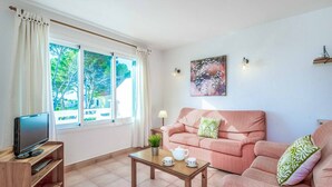 A living room with a TV on a wooden stand, a table with cups and a teapot, a pink sofa, and a window with curtains. - Villa Cati Punta Prima (Punta Prima)