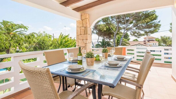 A covered outdoor dining area with a table set for two, surrounded by wicker chairs, a stone pillar, and a view of greenery and buildings in the background. - Villa Cati Punta Prima (Punta Prima)