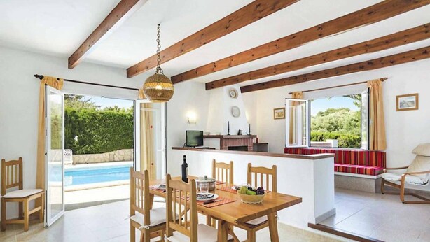A dining area with a wooden table and chairs, a kitchen area with a counter, and a view of a swimming pool. - Villa Bini Beatriz - Two Bedroom Villa, Sleeps 4 (Binibeca)