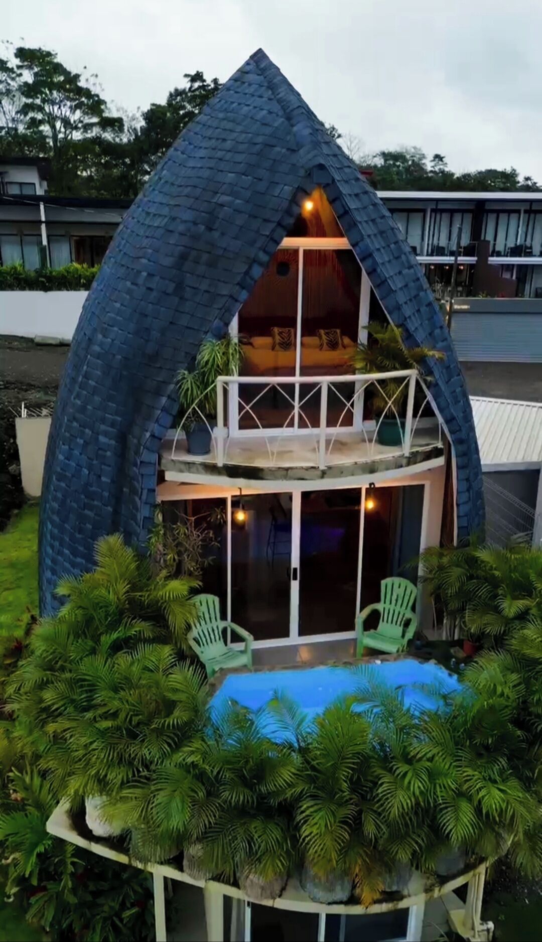 A two-story house with a steeply pitched roof, a balcony on the upper floor, and a pool area with green chairs.