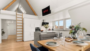 Een moderne keuken met een slaapverdieping, een eetgedeelte met een houten tafel gedekt voor een maaltijd, en een televisie aan de muur waarop 'NETFLIX' wordt weergegeven.