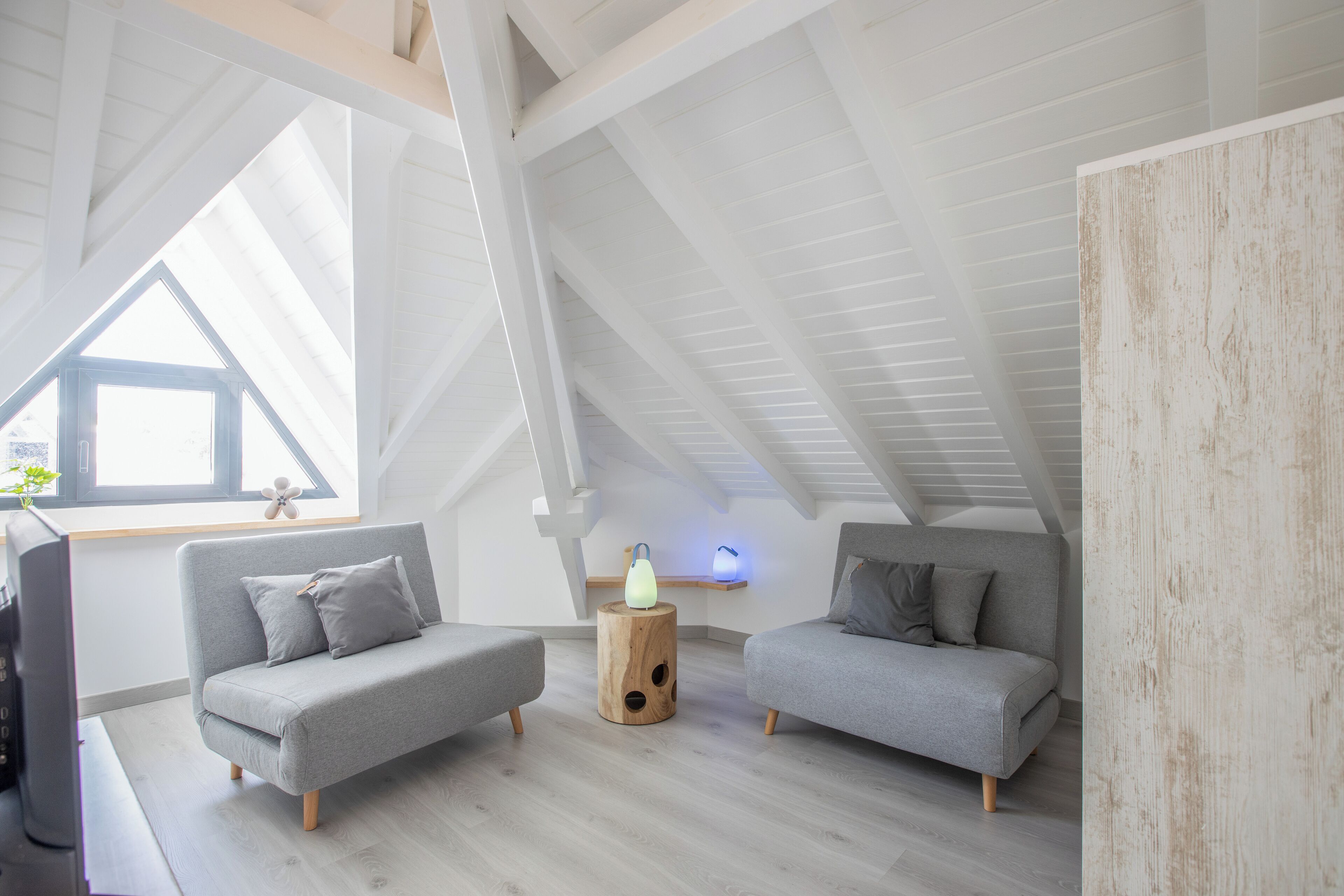 A modern loft bedroom with two grey sofas, a wooden side table, and a large window providing natural light.