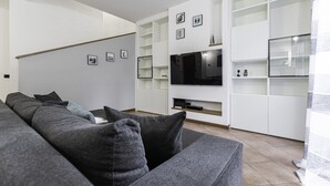 A modern living room with a grey sofa, a flat-screen TV mounted on a white wall, and built-in shelving with decorative items. - Villa Donatori (Lesmo)