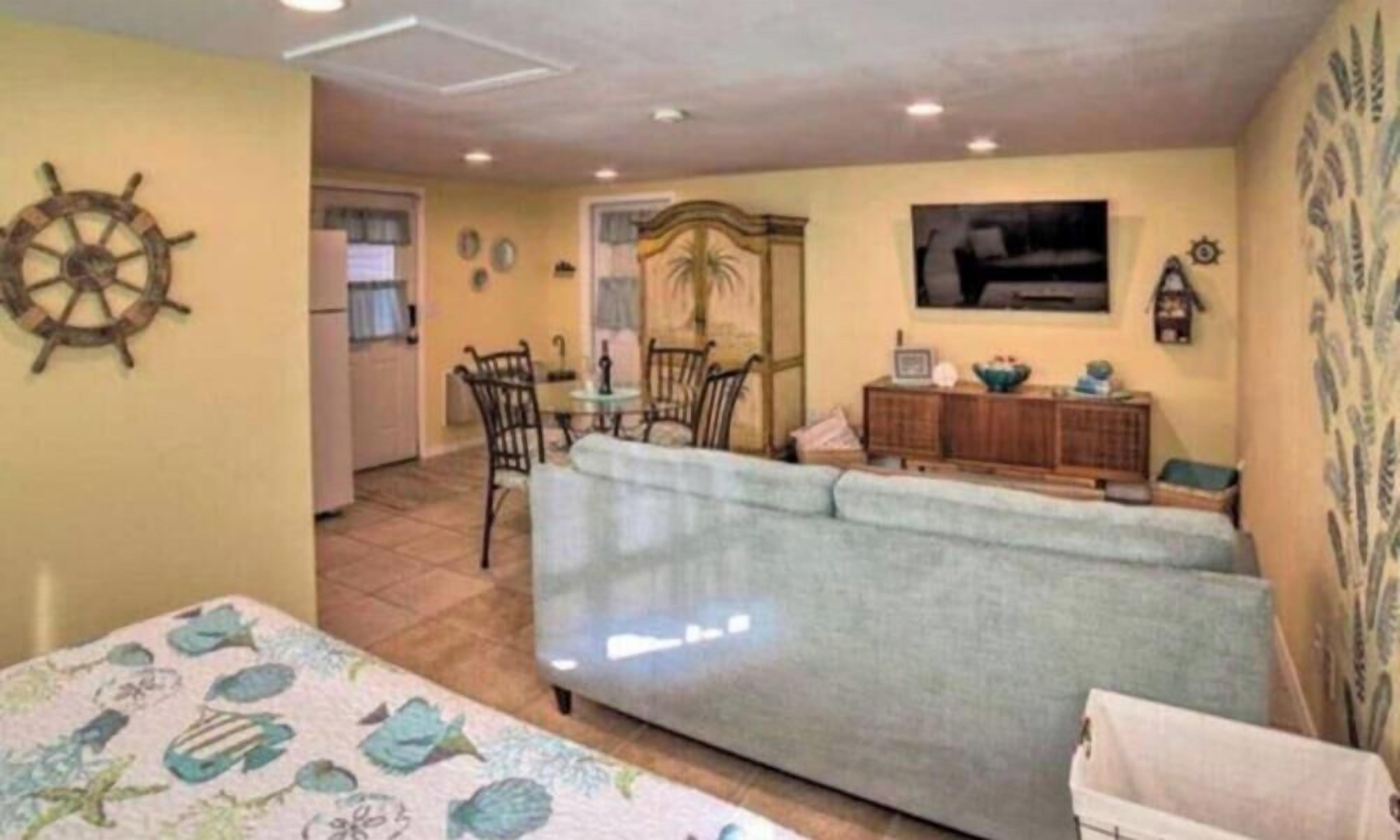 A living room with a sofa, a dining table with chairs, a kitchen area, and a decorative wall with a ship's wheel.