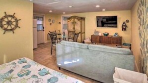 A living room with a sofa, a dining table with chairs, a kitchen area, and a decorative wall with a ship's wheel.
