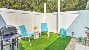 A backyard with a white fence, green artificial turf, two blue chairs, a small round table, and a barbecue grill.