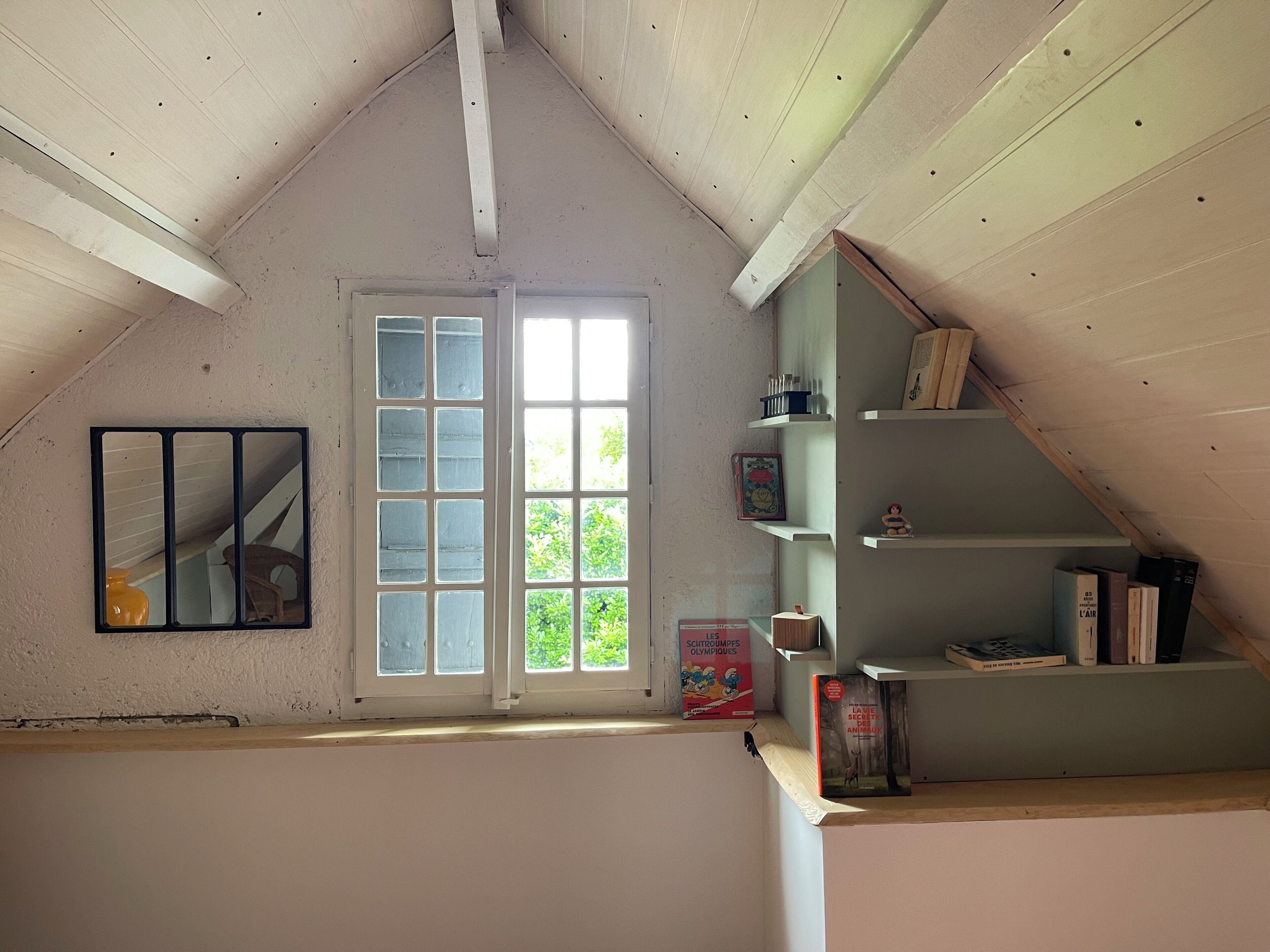 A room with a sloped ceiling, a large window with multiple panes, and a bookshelf with books and decorative items.