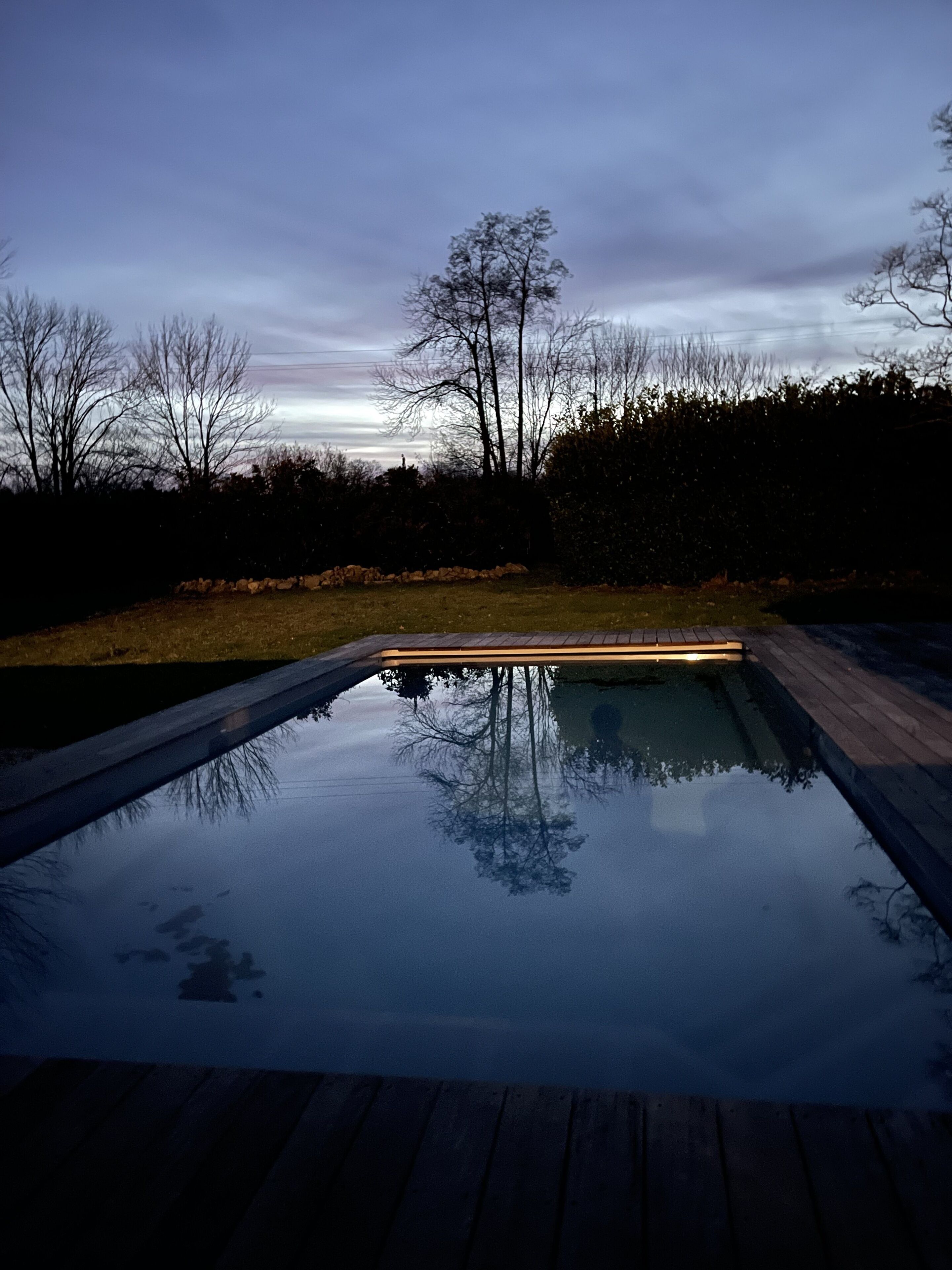 Piscina rectangular con un muelle de madera que la rodea, frente a un fondo de árboles y un seto.
