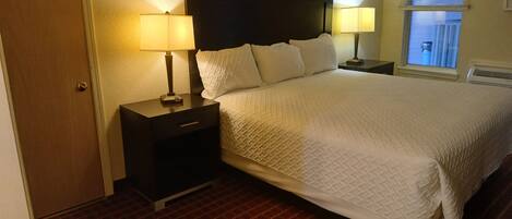 A hotel room with a large bed, two bedside tables with lamps, a window with blinds, and a door.