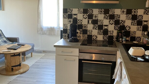 A modern kitchen with a built-in oven, a coffee maker, and a sink. There is a dining area with a round wooden table and a sofa.