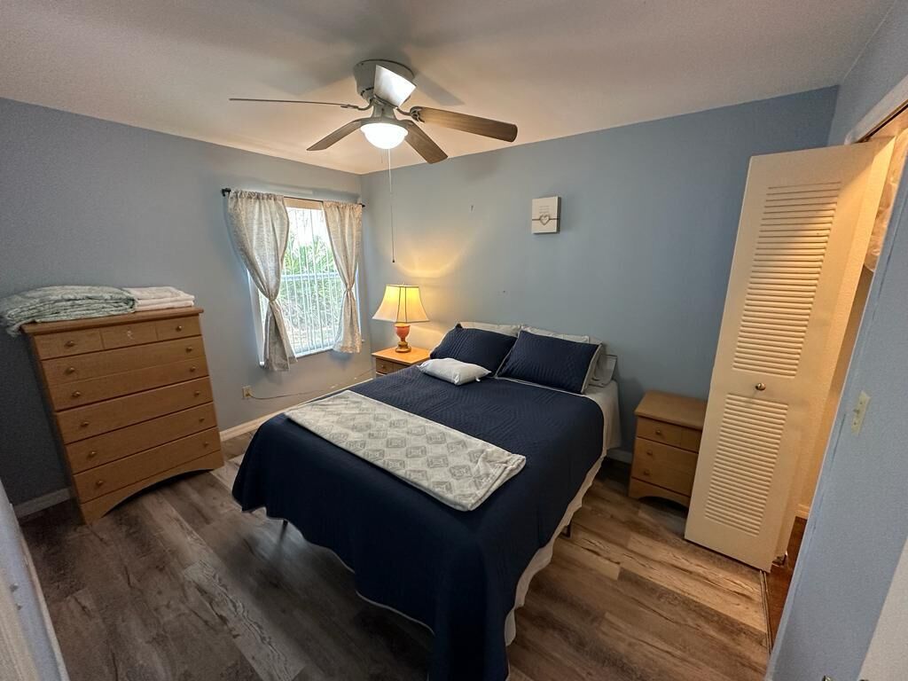 Quarto com cama, cómoda de madeira, mesas de cabeceira, ventoinha no teto, janela com cortinas e uma porta.