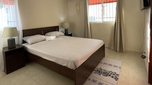 Un dormitorio con cama, mesitas de noche, lámpara, aire acondicionado, ventana con cortinas y televisor montado en la pared.