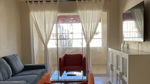 Una sala de estar con un sofá, una butaca roja, una mesa de vidrio, un ventilador de techo y un televisor en la pared.