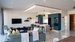 A modern living room with a grey sofa, a TV, and a kitchen area with blue cabinets and a bar stool. - The Bond Square - Two Bedroom Apartment, Sleeps 4 (Durban)