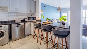 Cocina moderna con gabinetes blancos, microondas, lavavajillas y lavadora. Hay tres taburetes de bar y una pequeña isla con una planta.