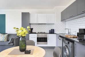 Cocina compacta con alacenas blancas, horno de acero inoxidable, lavadora y zona de comedor con mesa redonda y sillas.