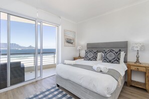 Een slaapkamer met een groot bed, een nachtkastje, een lamp en uitzicht op de oceaan door een schuifdeur.