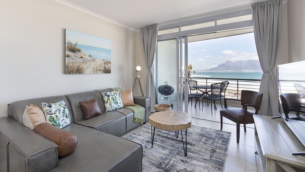 Un salon moderne avec un canapé gris, une table basse ronde et une vue sur l’océan grâce à de grandes fenêtres.