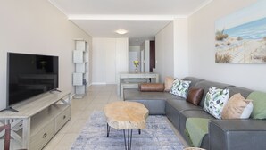 Un salon moderne avec un téléviseur à écran plat, un canapé gris agrémenté de plusieurs coussins, une table basse en bois et un tableau représentant une scène de plage accroché au mur.