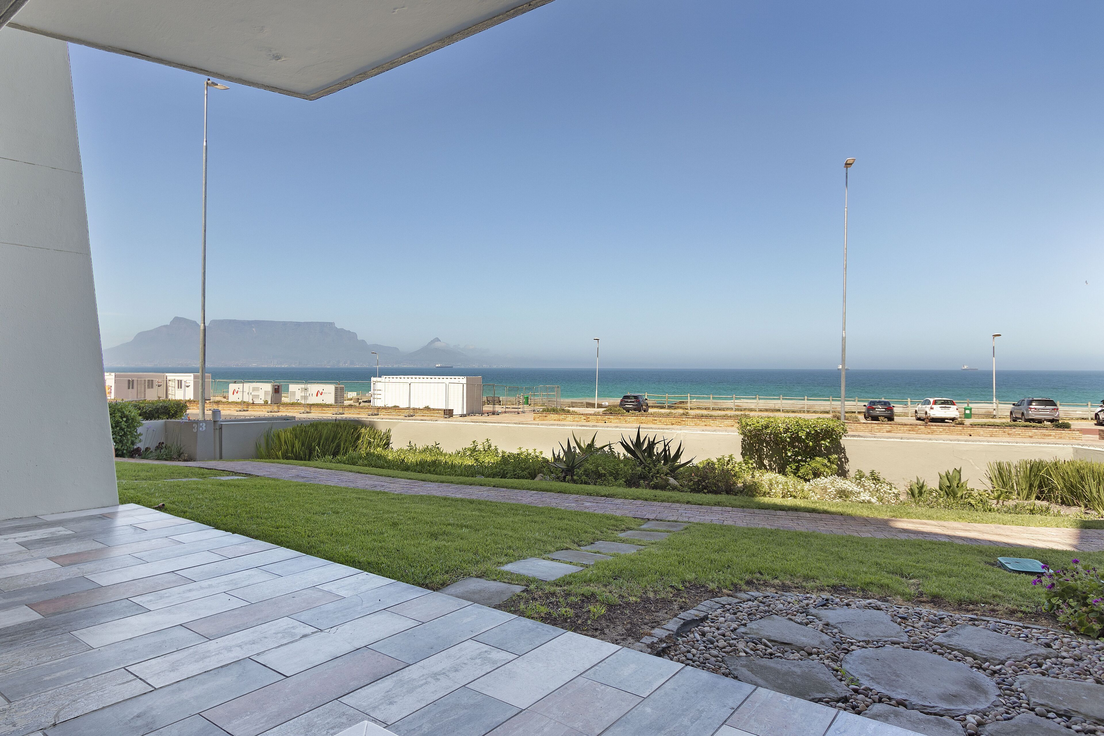 Une terrasse avec vue sur la plage et l’océan, un chemin pavé et un espace de stationnement avec des voitures.
