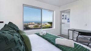 A modern hotel room with a large window offering a view of the sea and buildings, a neatly made bed with green and white bedding, and a small desk with a chair. - Sea Spray AG04 - Two Bedroom Apartment, Sleeps 4 (Cape Town)