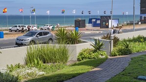 A coastal road with parked cars, a sidewalk with plants, and flags. - Sea Spray AG04 - Two Bedroom Apartment, Sleeps 4 (Cape Town)