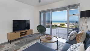 A modern living room with a flat-screen TV, a green chair, a sofa with patterned cushions, a wooden coffee table, and a view of the ocean through large glass doors. - Sea Spray AG04 - Two Bedroom Apartment, Sleeps 4 (Cape Town)