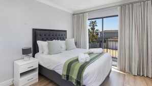 Una cama bien hecha con cabecera gris, ropa de cama blanca y una manta verde a rayas. Hay una mesita de noche blanca con una lámpara y un cajón. La habitación tiene un ventanal con cortinas y un balcón con vistas a palmeras y un edificio.