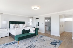 Quarto com cama, um banquinho, mesa de cabeceira, espelho e banheiro visível por uma porta aberta.
