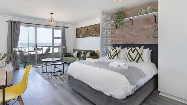 A modern hotel room with a large bed, a green sofa, a dining area, and a view of the sea. - Sea Spray B104 - Studio Apartment, Sleeps 2 (Cape Town)