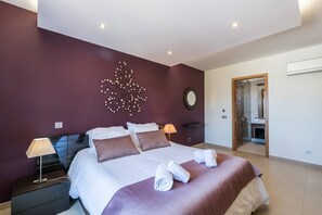 A modern bedroom with a large bed, two bedside lamps, a decorative wall piece, and a bathroom visible through an open door. - Captivating 2-bed Apartment in Praia da Luz (Lagos)