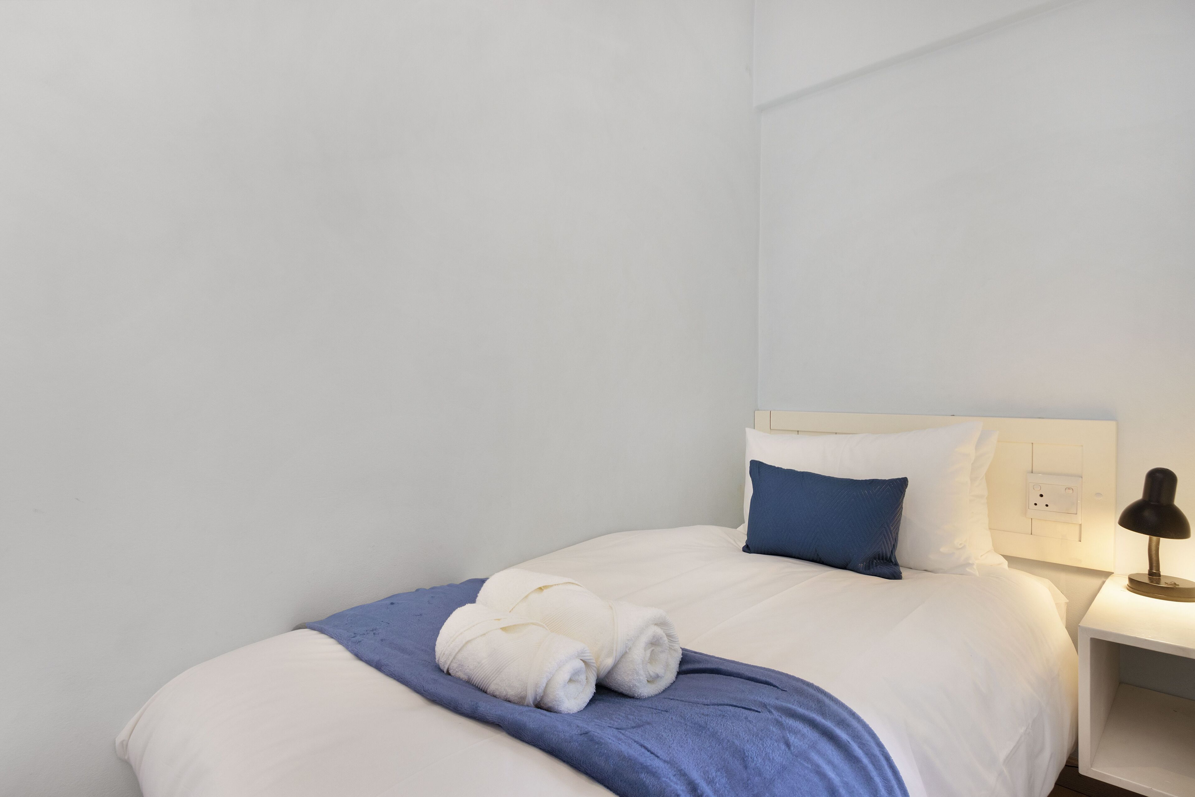 A neatly made bed with white bedding and a blue throw blanket, a single blue pillow, and a small bedside table with a lamp.