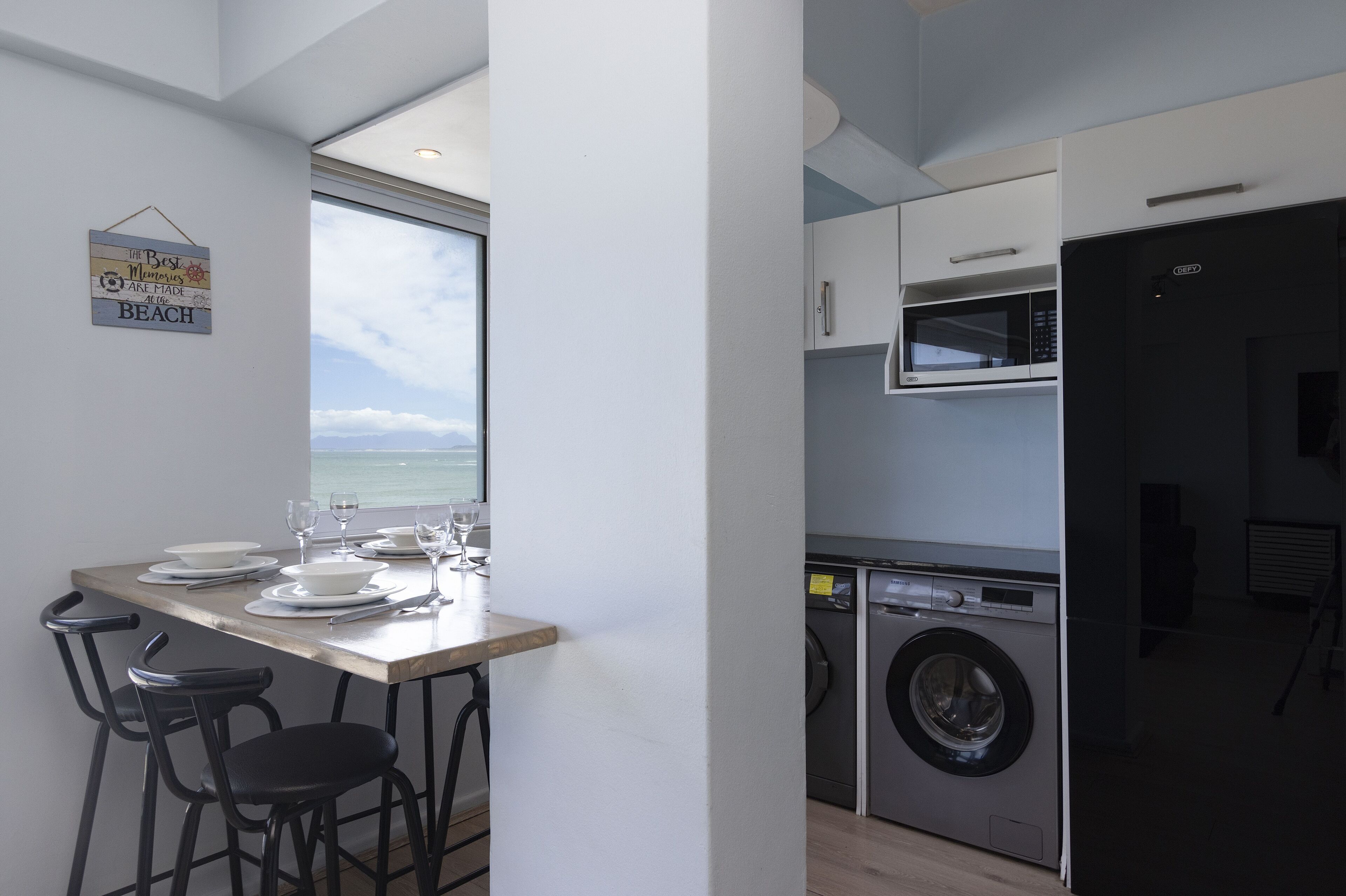 A compact kitchen with a view of the ocean, a washing machine, and a dining area with chairs.