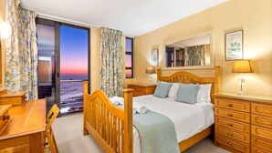 Een hotelkamer met een houten bed, een bureau, een dressoir en uitzicht op de ondergaande zon door het raam.