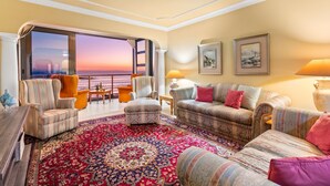 Een ruime woonkamer met een groot raam dat uitzicht biedt op een zonsondergang, comfortabele zitplaatsen en een decoratief vloerkleed.