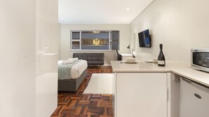 A modern hotel room with a bed, a small kitchenette, a flat-screen TV mounted on the wall, and a view of the city through the window. - Sandringham 55 - Studio Apartment, Sleeps 2 (Cape Town)