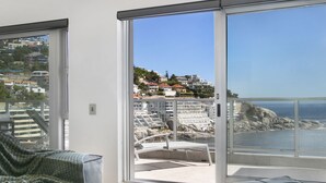 A balcony with a view of a coastal town, a white chair, and a grey cushioned bench. - Seacliffe 502 - Two Bedroom Apartment, Sleeps 4 (Cape Town)