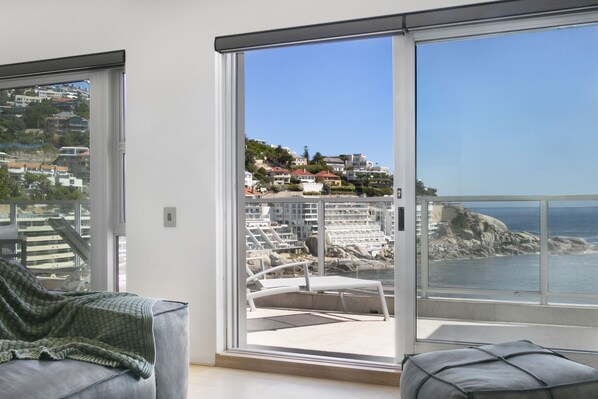A room with a large sliding glass door leading to a balcony with a view of a coastal area and buildings.