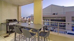 An outdoor balcony with a grill, table, and chairs, overlooking a building with a sign reading "S.W.A.T."