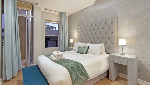 A hotel room with a large bed, two bedside tables with lamps, a green throw blanket, and a view of the outside through a sliding door.