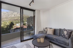 A modern living room with a grey sofa, two cushions, a small round table, and a view of a mountainous landscape through large glass doors.