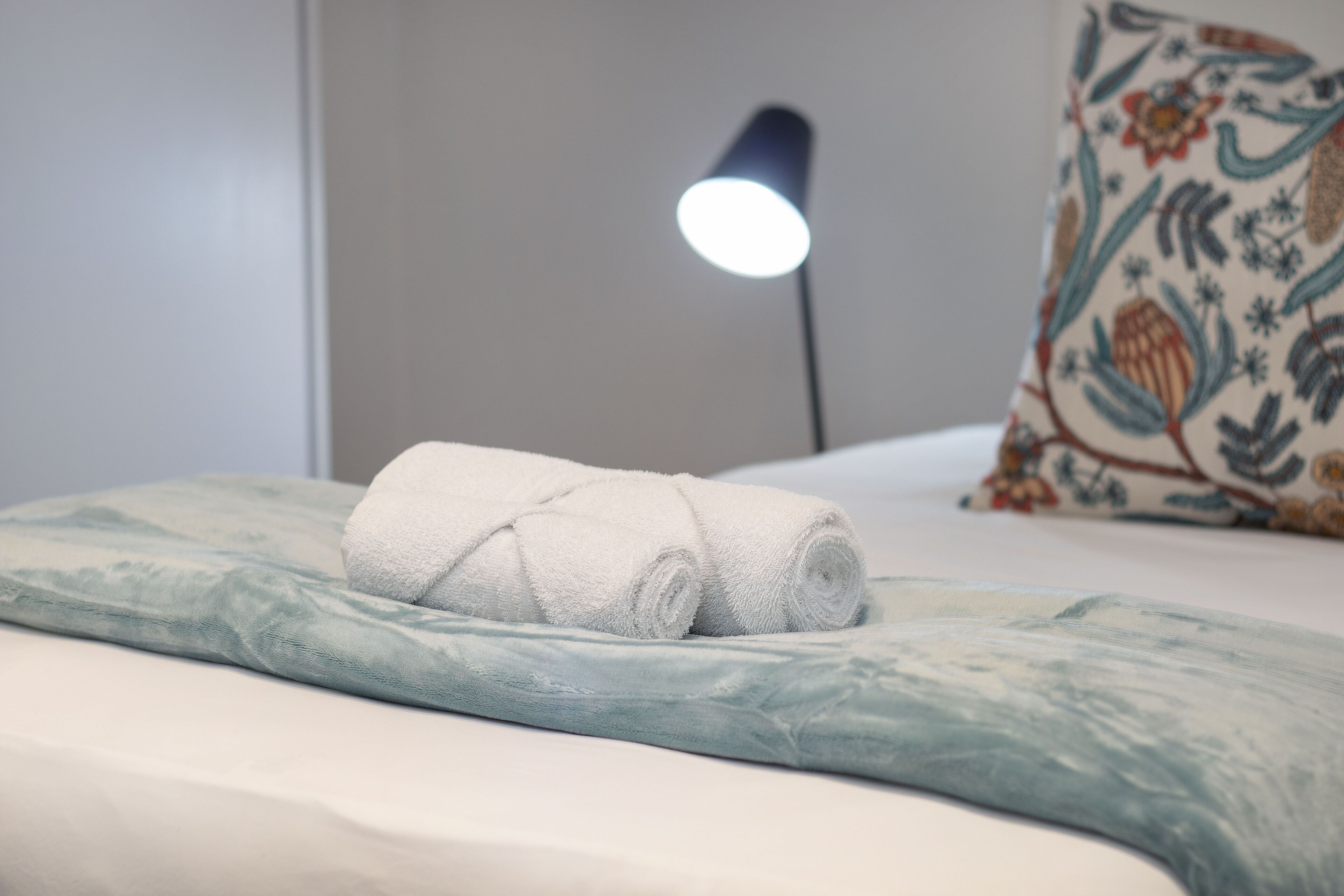 Una cama bien hecha con una toalla blanca enrollada y una almohada azul claro.