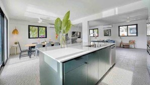 A modern kitchen with a central island, a dining area with a table and chairs, and a large window providing natural light. - Purple Palace -Fully Renovated w/Pool & Ocean View (Rincón)